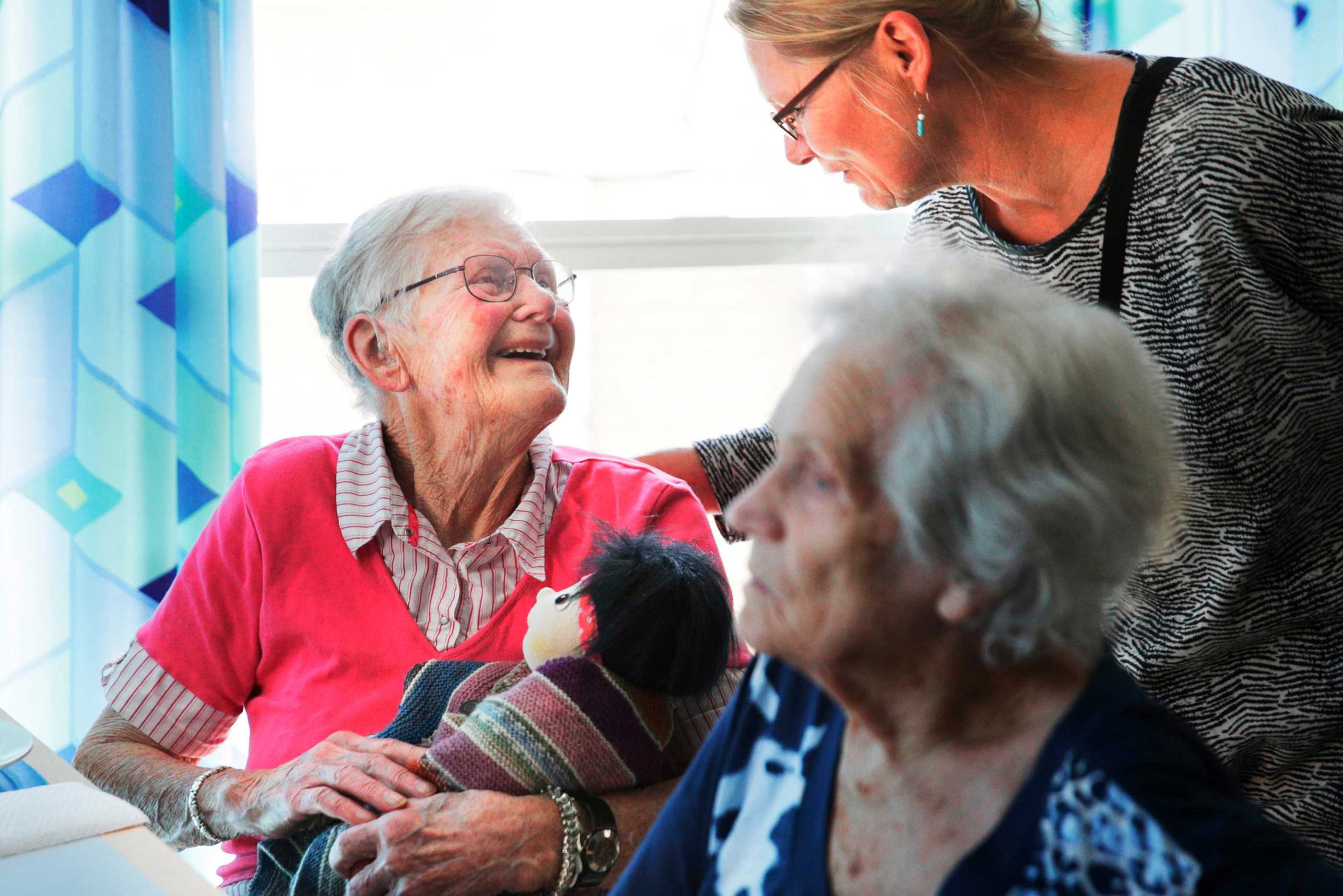 Dame der sidder med dukke kigger smilende op til dame der kigger ned på hende